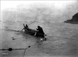 Two people in a canoe pulling a fishing net. A bluff above the ocean in the background. Image is black and white. 